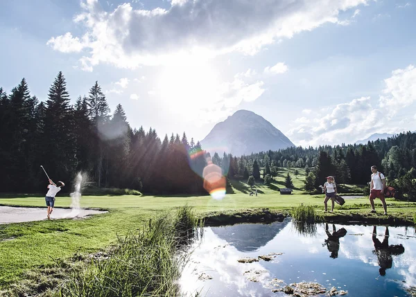 Person spielt Golf auf grünem Golfplatz mit Bergen und Wald im Hintergrund