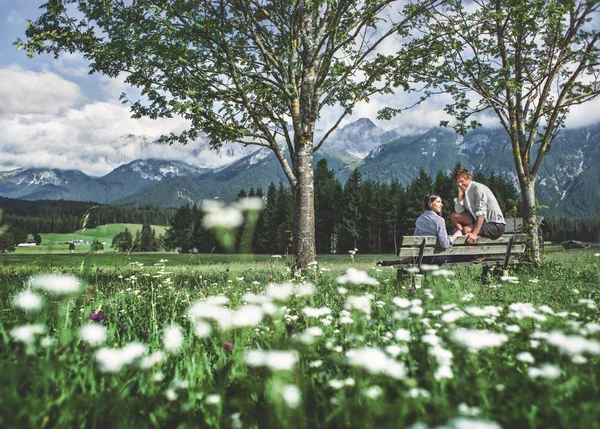 Zwei Personen sitzen auf einer Bank in blumiger Bergwiese mit Alpen im Hintergrund