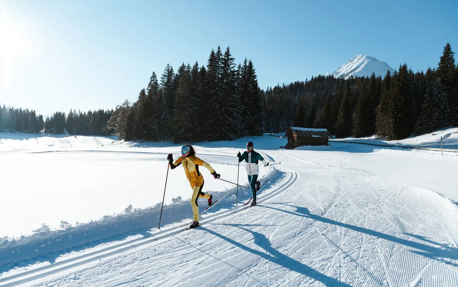 Zwei Langläufer laufen auf einer sonnigen Schneepiste in den Bergen