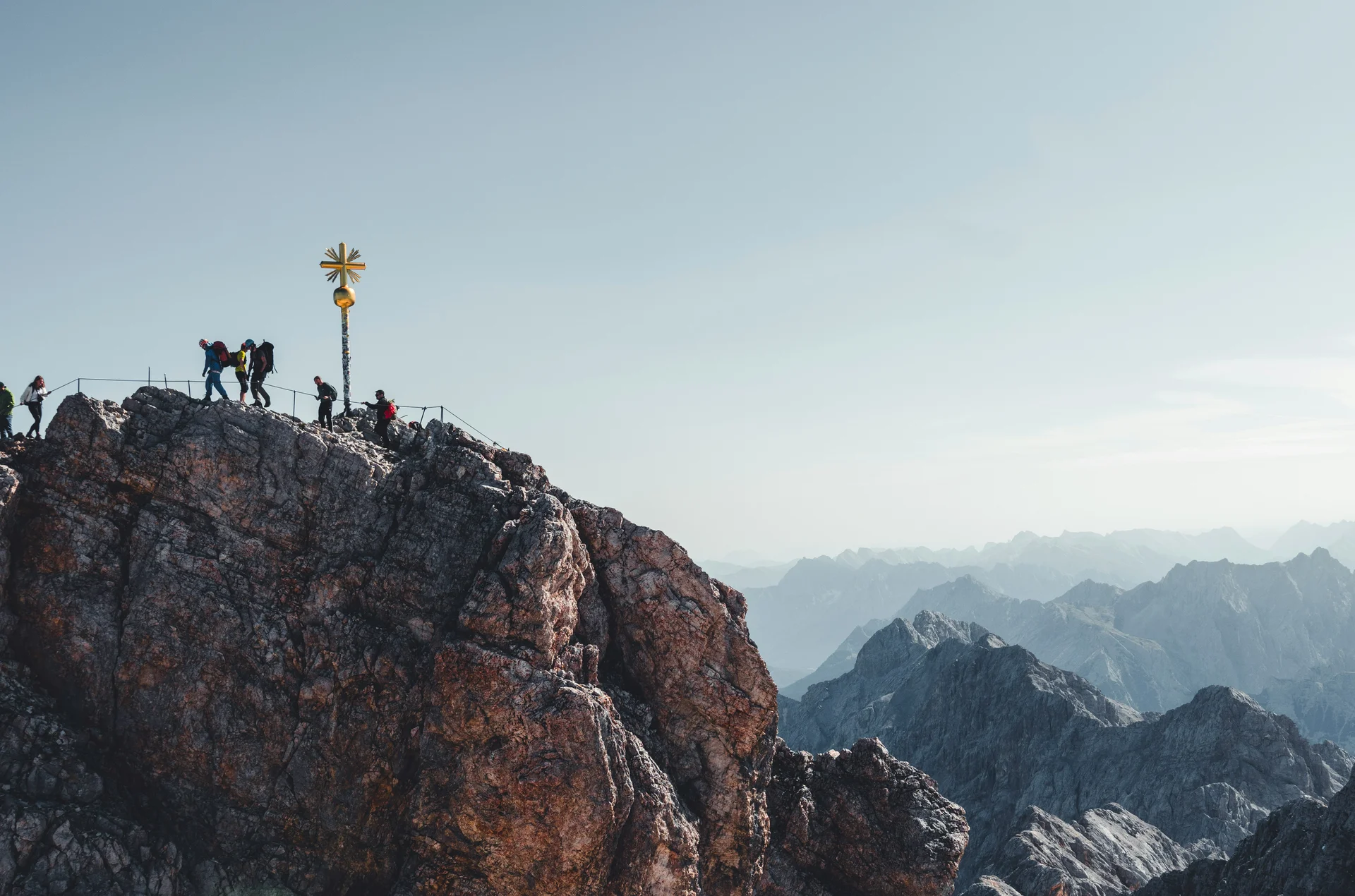 Wanderer auf felsigem Berggipfel mit Gipfelkreuz bei klarem Himmel