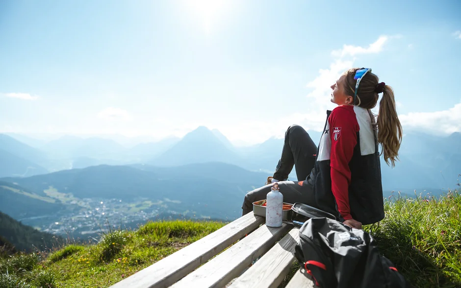 Erholung oder Regeneration? Warum viele Menschen nach dem Urlaub wieder müde sind Frau sitzt auf Bank mit Bergblick bei sonnigem Wetter