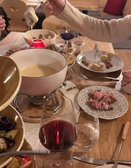 Person taucht Brot in Käsesfondue an gedecktem Holztisch mit Wein und Fleisch