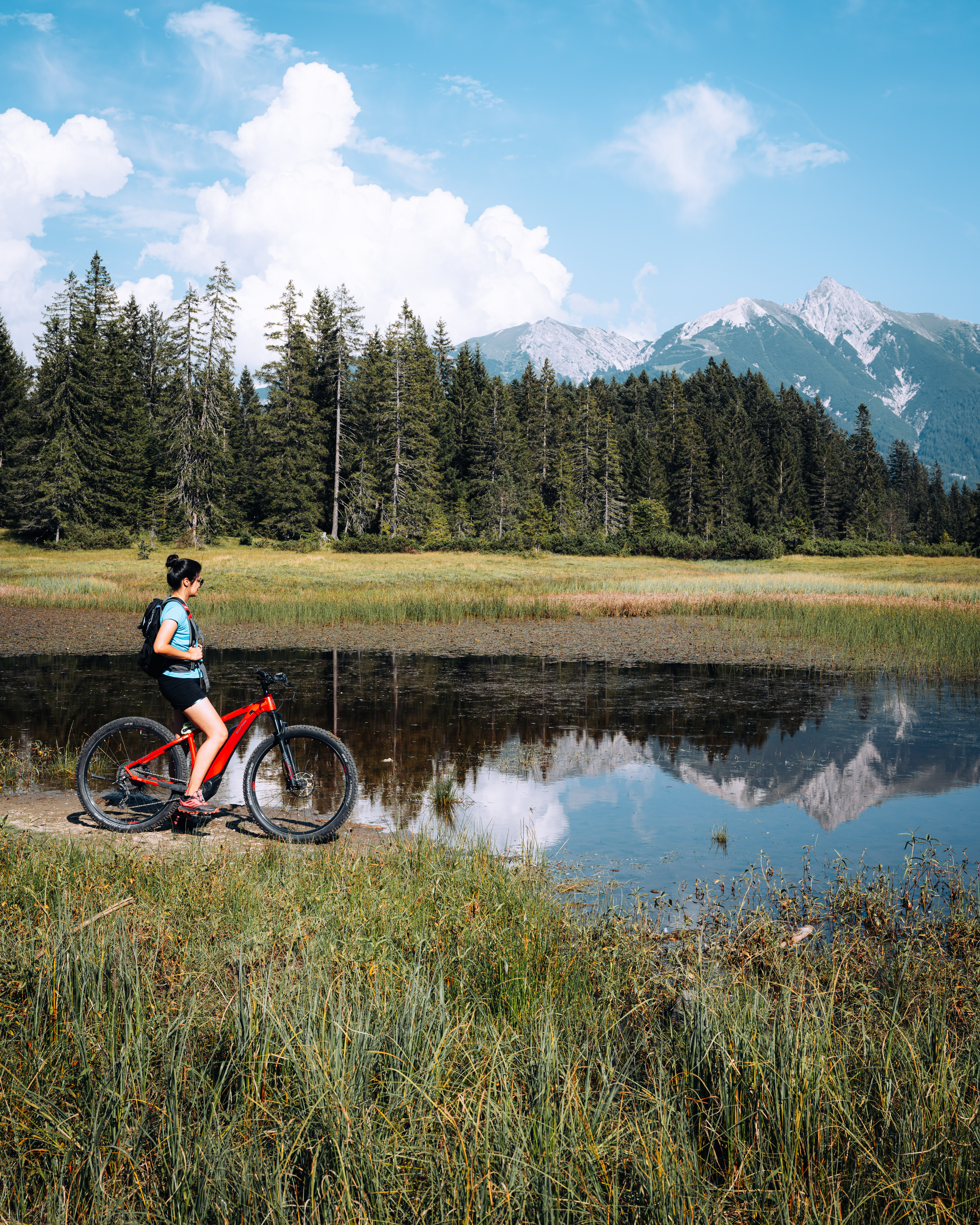 Werfen Sie einen Blick in die Bildergalerie Frau mit rotem Fahrrad am See vor Wald und Bergen bei klarem Himmel